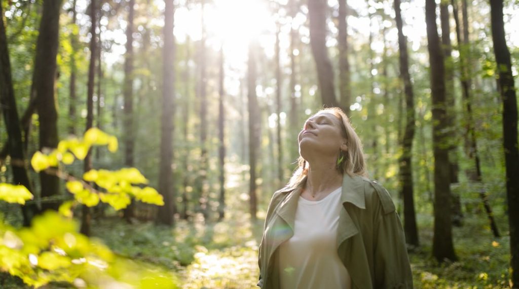 Claudia Keil-Werner steht entspannt in einem sonnendurchfluteten Wald, schließt die Augen und atmet tief ein. Die Sonnenstrahlen fallen durch die hohen Bäume und erzeugen eine friedliche, naturverbundene Atmosphäre.
