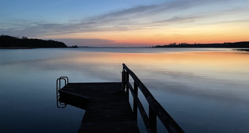 Ein Steg mit einer Badeleiter führt in einen ruhigen See, der das farbenprächtige Licht eines Sonnenuntergangs widerspiegelt. Der Himmel zeigt einen Verlauf von Blau zu Orange und Rosa. Am Horizont sind dunkle Landstriche zu sehen.